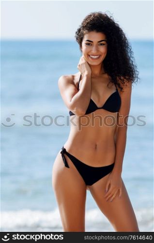 Young arabic woman with beautiful body in swimwear smiling in a tropical beach. Brunette female with curly long hairstyle wearing black bikini.. Young arabic woman with beautiful body in swimwear smiling on a tropical beach.