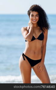 Young arabic woman with beautiful body in swimwear smiling in a tropical beach. Brunette female with curly long hairstyle wearing black bikini.. Young arabic woman with beautiful body in swimwear smiling on a tropical beach.