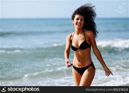 Young arabic woman with beautiful body in swimwear smiling on a . Young arabic woman with beautiful body in swimwear smiling in a tropical beach. Brunette female with curly long hairstyle wearing black bikini.