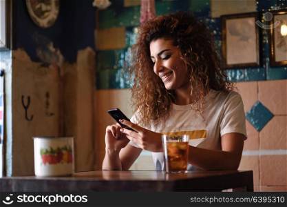 Young arabic woman with black curly hairstyle sitting in a beautiful bar with vintage decoration. Arab girl in casual clothes drinking a soda and looking at her smart phone