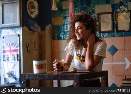 Young arabic woman with thoughtful gesture sitting in a beautiful bar with vintage decoration. Arab girl in casual clothes drinking a soda.. Arabic woman in a beautiful bar looking through the window