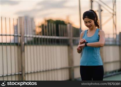 Young Asia athlete lady exercises checking progress looking heart rate monitor on smartwatch in urban city. Teen girl runner resting exhausted after intense running workout training cardio in morning.