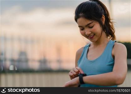 Young Asia athlete lady exercises checking progress looking heart rate monitor on smartwatch in urban city. Teen girl runner resting exhausted after intense running workout training cardio in morning.