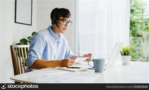 Young Asia businessman using laptop talk to colleagues about plan in video call while smart working from home at living room. Self-isolation, social distancing, quarantine for corona virus prevention.