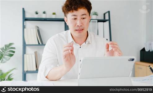 Young Asia businessman using tablet talk to colleagues about plan in video call while smart working from home at living room. Self-isolation, social distancing, quarantine for corona virus prevention.