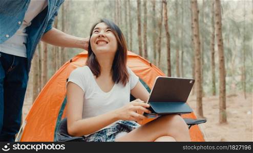 Young asia campers couple using tablet in chairs by tent in forest. Male and female traveler relaxing and talking on a summer day at campsite. Outdoor activity, adventure travel, or holiday vacation.