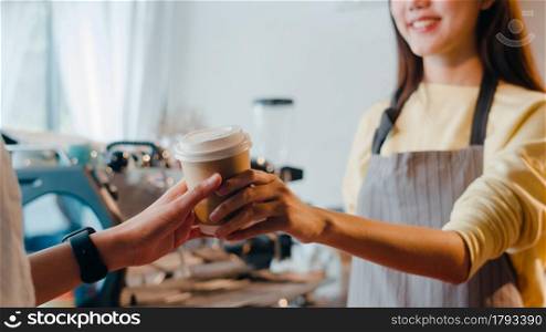 Young Asia female barista serving take away hot coffee paper cup to consumer standing behind bar counter at cafe restaurant. Owner small business, food and drink, service mind concept.
