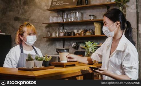 Young Asia female barista wear face mask serving take away hot coffee paper cup to consumer at cafe. Owner small business, lifestyle new normal after corona virus and social distancing concept.