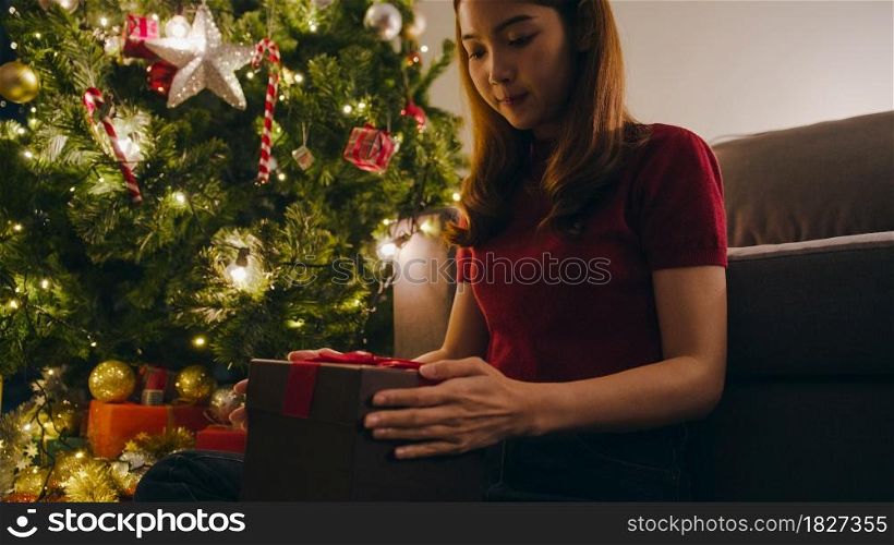 Young Asia female having fun opening X&rsquo;Mas present box near Christmas tree decorated with ornaments in living room at home. Merry Christmas night and Happy New Year holiday festival.