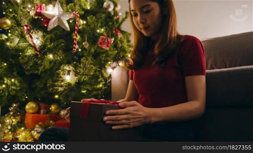 Young Asia female having fun opening X&rsquo;Mas present box near Christmas tree decorated with ornaments in living room at home. Merry Christmas night and Happy New Year holiday festival.