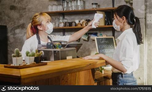 Young Asia female restaurant staff wearing protective face mask using infrared thermometer checker or temperature gun on customer’s forehead before enter. Lifestyle new normal after corona virus.