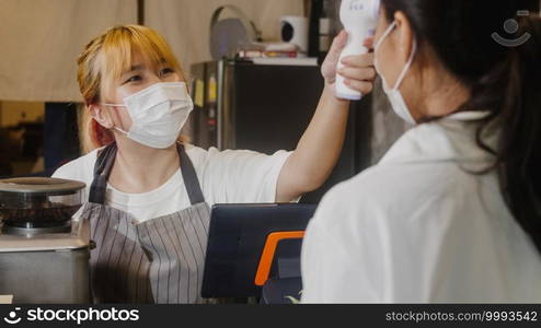 Young Asia female restaurant staff wearing protective face mask using infrared thermometer checker or temperature gun on customer’s forehead before enter. Lifestyle new normal after corona virus.