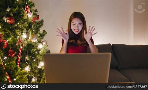 Young Asia female using laptop video call talking with couple with X&rsquo;Mas present box, Christmas tree decorated with ornament in living room at home. Christmas night and New Year holiday festival.