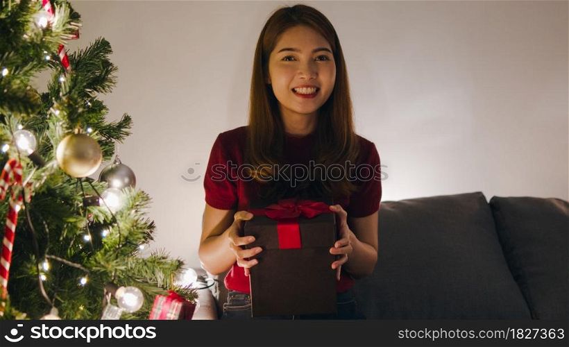Young Asia female using mobile phone video call talking with couple with X&rsquo;Mas present box, Christmas tree decorated with ornament in living room at home. Christmas night and New Year holiday festival