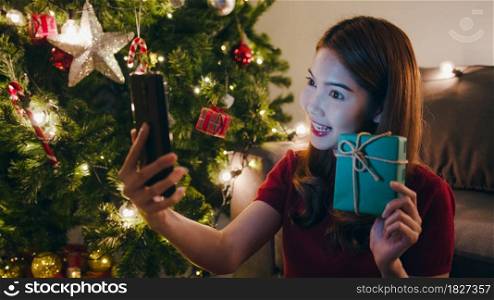 Young Asia female using smart phone video call talking with couple with X&rsquo;Mas present box, Christmas tree decorated with ornament in living room at home. Christmas night and New Year holiday festival.