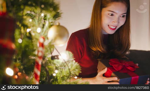 Young Asia female using tablet video call talking with couple with X&rsquo;Mas present box, Christmas tree decorated with ornament in living room at home. Christmas night and New Year holiday festival.