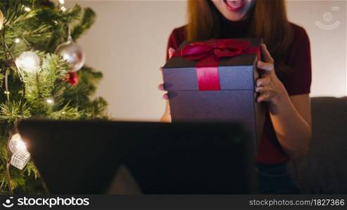 Young Asia female using tablet video call talking with couple with X&rsquo;Mas present box, Christmas tree decorated with ornament in living room at home. Christmas night and New Year holiday festival.