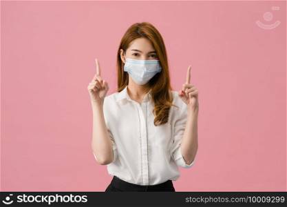 Young Asia girl wearing medical face mask shows something at blank space with dressed in casual cloth and looking at camera isolated on pink background. Social distancing, quarantine for corona virus.