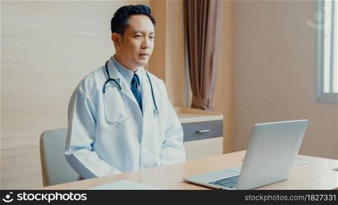 Young Asia guy doctor in white medical uniform with stethoscope using computer laptop talking video conference call with patient at desk in health clinic or hospital. Consulting and therapy concept.