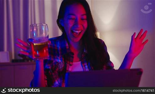 Young Asia lady drinking beer having fun happy moment disco neon night party event online celebration via video call in living room at home. Social distancing, quarantine for coronavirus prevention.