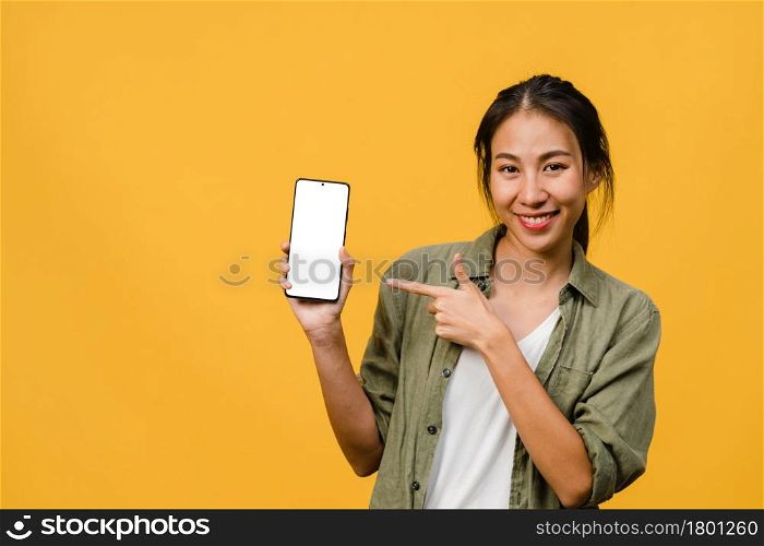 Young Asia lady show empty smartphone screen with positive expression, smiles broadly, dressed in casual clothing feeling happiness on yellow background. Mobile phone with white screen in female hand.