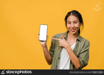 Young Asia lady show empty smartphone screen with positive expression, smiles broadly, dressed in casual clothing feeling happiness on yellow background. Mobile phone with white screen in female hand.