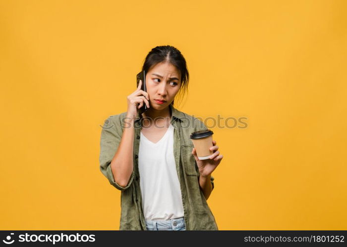 Young Asia lady talk by phone and hold coffee cup with negative expression, excited screaming, cry emotional angry in casual cloth and stand isolated on yellow background. Facial expression concept.