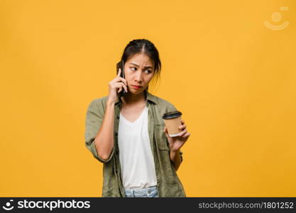 Young Asia lady talk by phone and hold coffee cup with negative expression, excited screaming, cry emotional angry in casual cloth and stand isolated on yellow background. Facial expression concept.