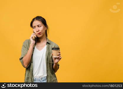 Young Asia lady talk by phone and hold coffee cup with negative expression, excited screaming, cry emotional angry in casual cloth and stand isolated on yellow background. Facial expression concept.