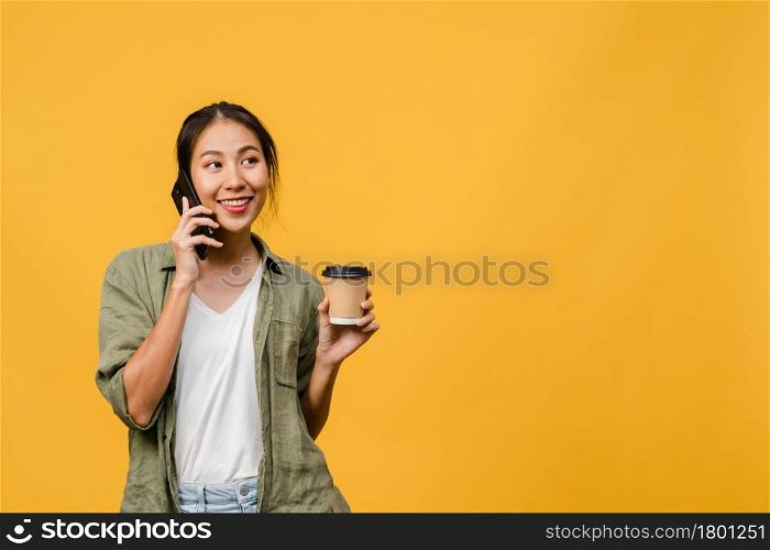 Young Asia lady talk by phone and hold coffee cup with positive expression, smile broadly, dressed in casual cloth feeling happiness and stand isolated on yellow background. Facial expression concept.