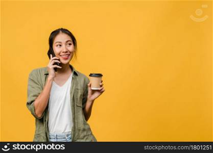 Young Asia lady talk by phone and hold coffee cup with positive expression, smile broadly, dressed in casual cloth feeling happiness and stand isolated on yellow background. Facial expression concept.