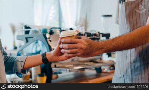 Young Asia male barista serving take away hot coffee paper cup to consumer standing behind bar counter at cafe restaurant. Owner small business, food and drink, service mind concept.
