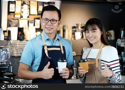 Young asia man and woman barista holding a disposable coffee cup and thumb up with smiling face at cafe counter background, small business owner, food and drink industry