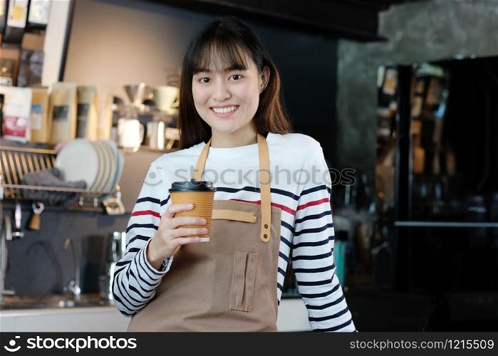 Young asia woman barista holding a disposable coffee cup with smiling face at cafe counter background, small business owner, food and drink industry