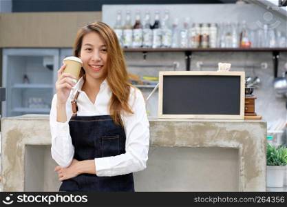 Young asia woman barista holding a disposable coffee cup with smiling face at cafe counter background, small business owner, food and drink industry concept