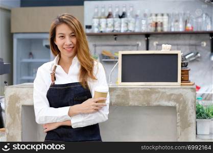 Young asia woman barista holding a disposable coffee cup with smiling face at cafe counter background, small business owner, food and drink industry concept