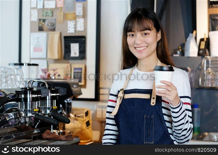 Young asia woman barista holding a disposable coffee cup with smiling face at cafe counter background, small business owner, food and drink industry