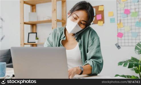 Young Asia women wear medical face mask talking on phone busy entrepreneur working distantly in living room. Working from home, remotely work, social distancing, quarantine for corona virus prevention