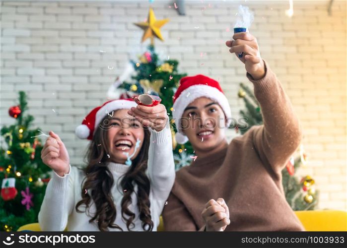 Young asian adult teenager couple celebrateing christmas holiday together with Firecrackers in living room with christmas tree decoration. (Selective Focus on Firecracker)