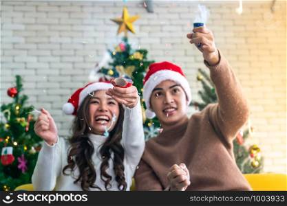 Young asian adult teenager couple celebrateing christmas holiday together with Firecrackers in living room with christmas tree decoration. (Selective Focus on Firecracker)