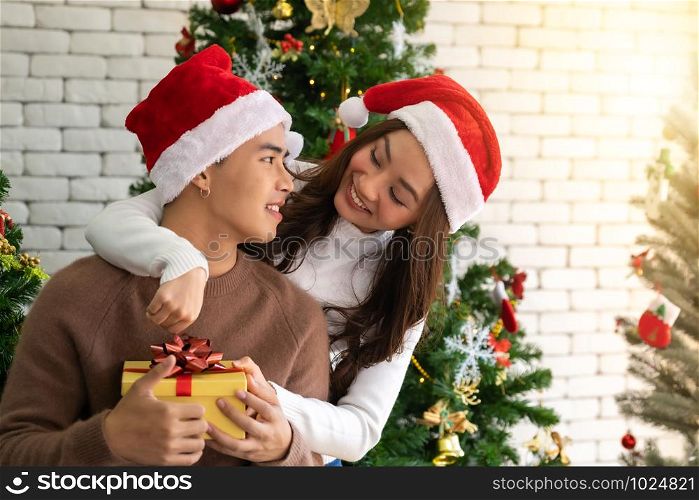 Young asian adult teenager couple holding gift present box hugging and celebrateing christmas holiday together in living room with christmas tree ornament decoration.