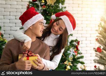 Young asian adult teenager couple holding gift present box hugging and celebrateing christmas holiday together in living room with christmas tree ornament decoration.