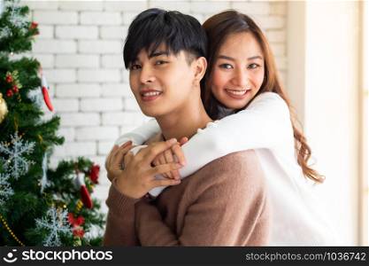 Young asian adult teenager couple hugging and celebrateing christmas holiday together in living room with christmas tree ornament decoration.