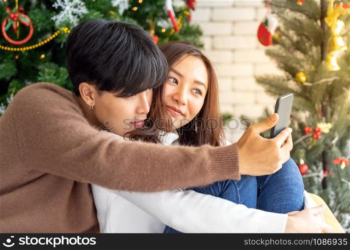 Young asian adult teenager couple make thier selfie photographing for celebrateing christmas holiday together in living room with christmas tree decoration.