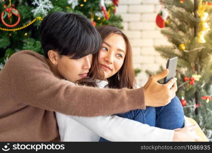 Young asian adult teenager couple make thier selfie photographing for celebrateing christmas holiday together in living room with christmas tree decoration.