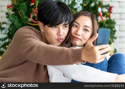 Young asian adult teenager couple make thier selfie photographing for celebrateing christmas holiday together in living room with christmas tree decoration.