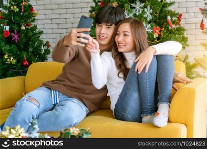 Young asian adult teenager couple take selfie photographing for celebrateing christmas holiday together in living room with christmas tree decoration.