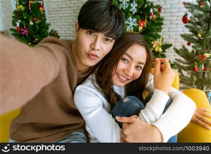 Young asian adult teenager coupletake selfie photographing for celebrateing christmas holiday together in living room with christmas tree decoration.