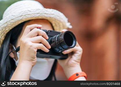 Young asian backpacker female wearing hat and protection mask while traveling in historic site, she use camera take a photo with happy