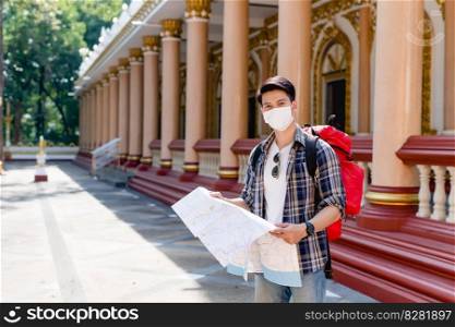 Young Asian backpacker male in face mask standing and checking direction on paper map in hand at beautiful Thai temple, looking at camera 